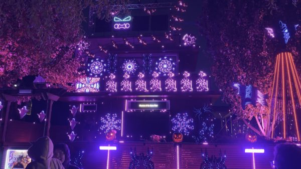 The house in purple light with pixel snowflake spinners, ghost props, jack-o'-lanterns along the fence, and the mega tree